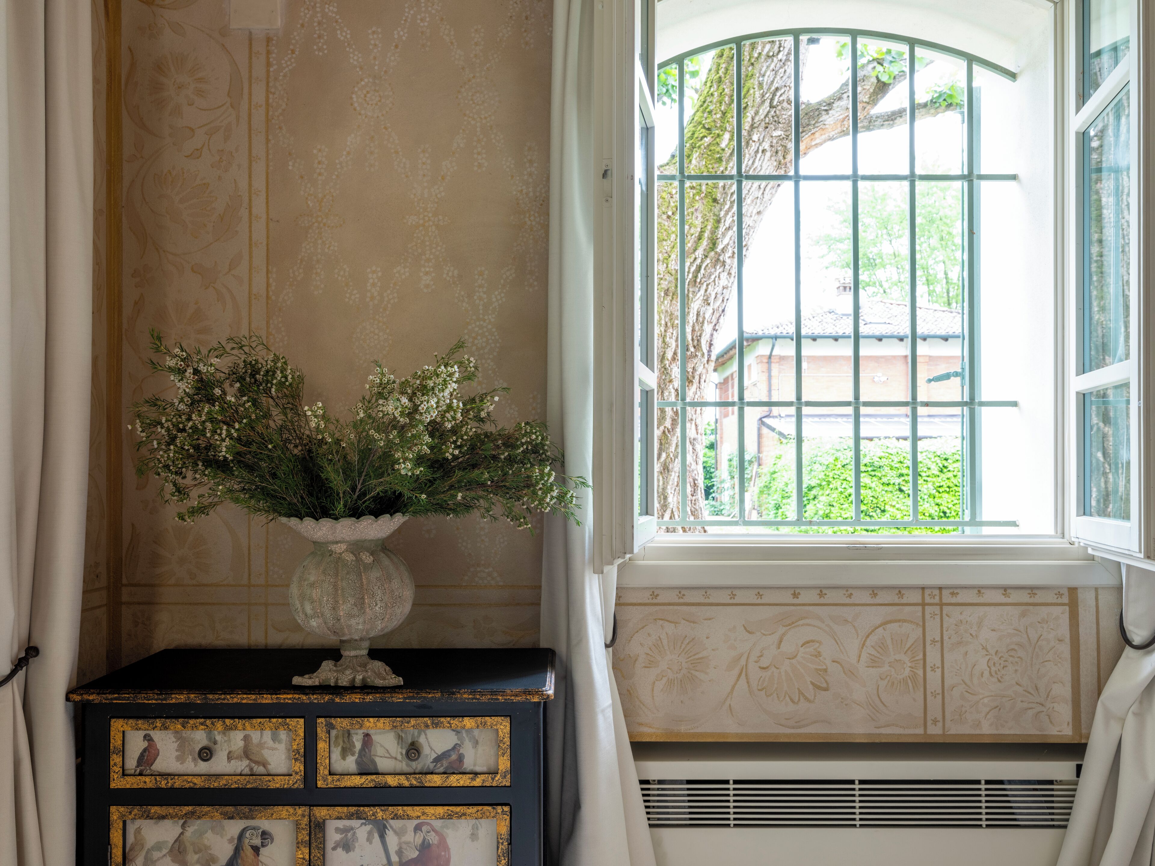 A room with a window, a decorative vase with flowers, and a dresser with carved panels.