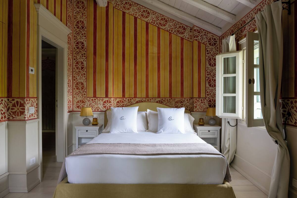 A hotel room with a large bed, two bedside tables with lamps, a window with curtains, and a door leading to another room.