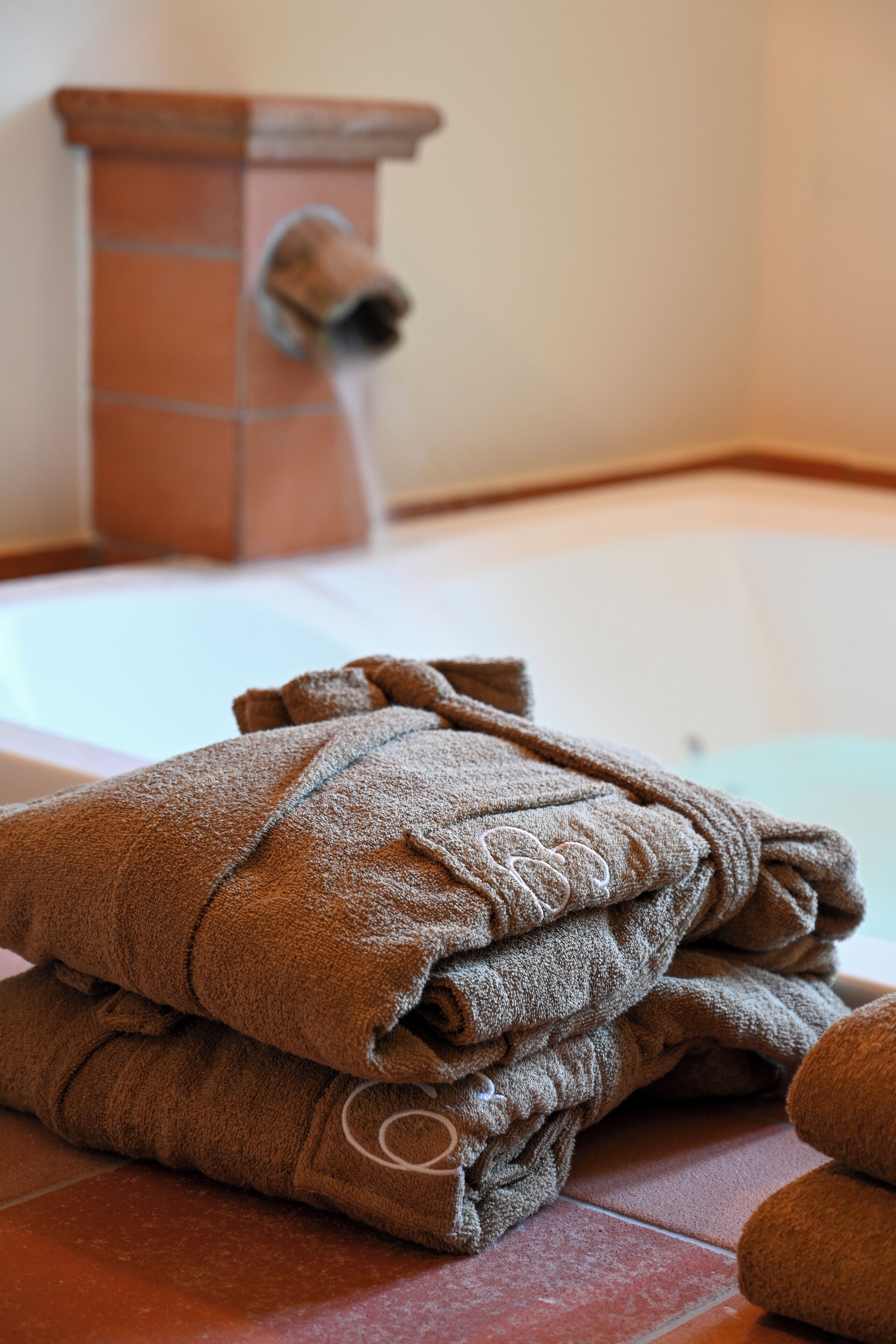 A stack of brown towels on a tiled floor with a bathtub and a terracotta-colored object in the background.
