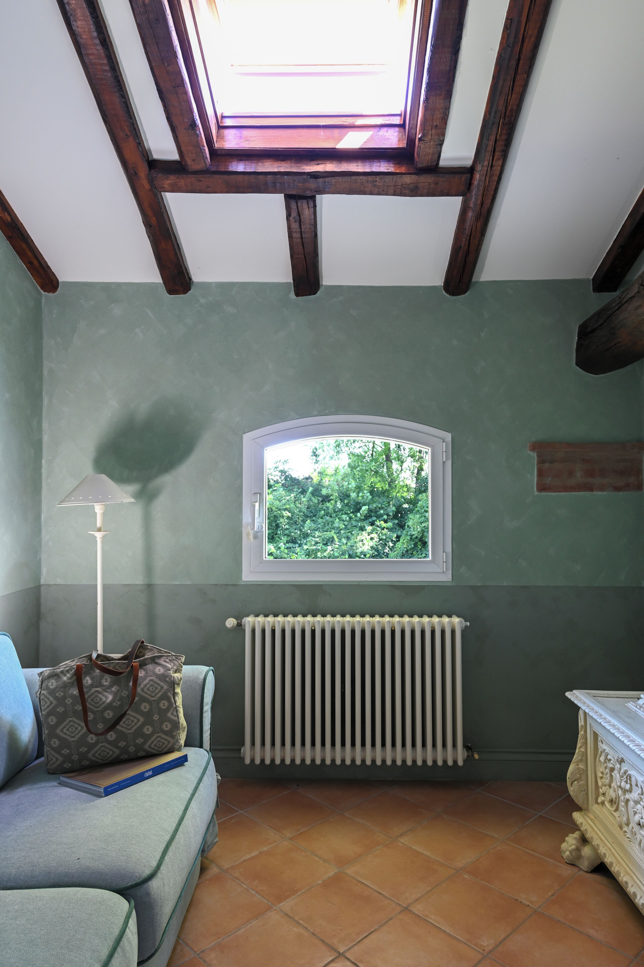 Una habitación con tragaluz, un radiador, una ventana con vistas a zonas verdes, un sofá y una lámpara.