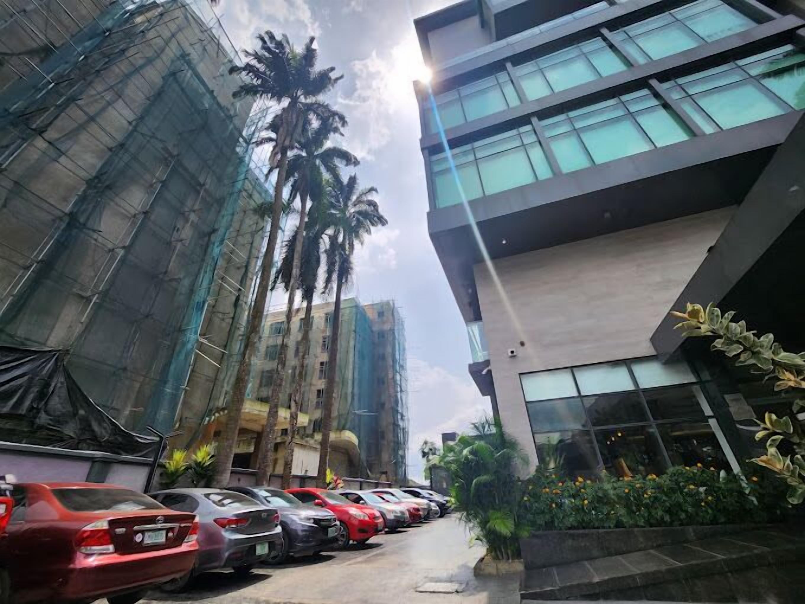 Un estacionamiento con varios autos, un edificio moderno con ventanas amplias y palmeras al fondo.