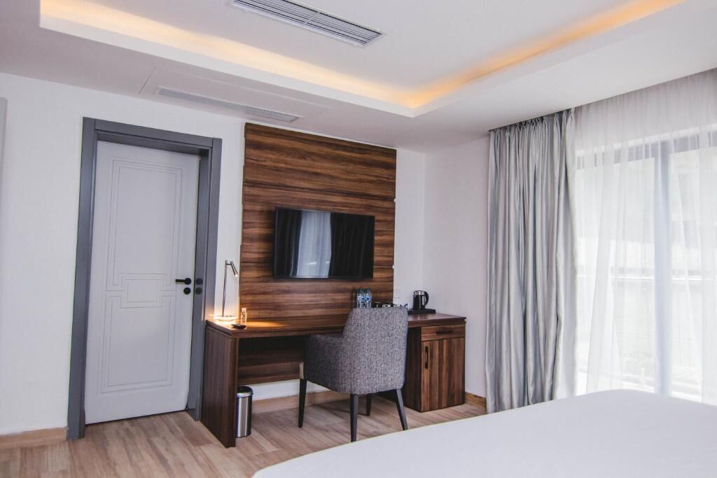A hotel room with a wooden accent wall, a flat-screen TV, a desk with a chair, a bed, and a large window with curtains.