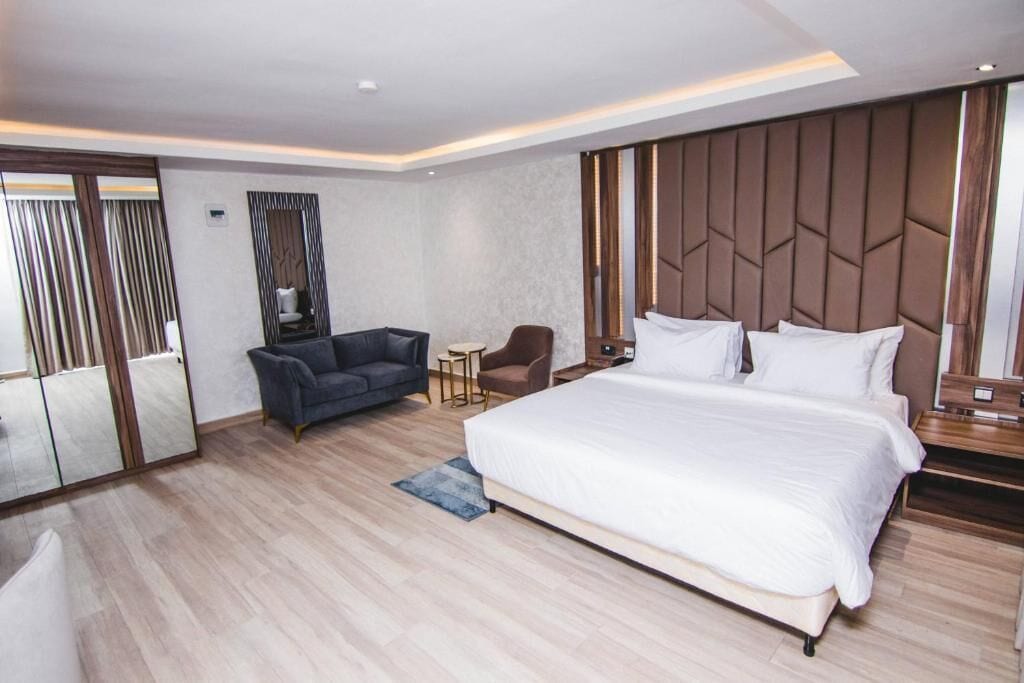 A modern hotel room with a large bed, a sofa, a chair, a small table, and a mirror.