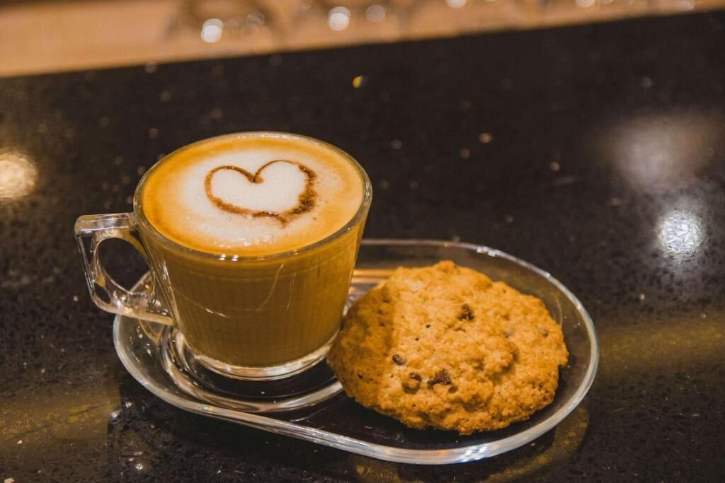 A clear glass cup of coffee with a heart-shaped latte art on a saucer, accompanied by a cookie on a small plate.