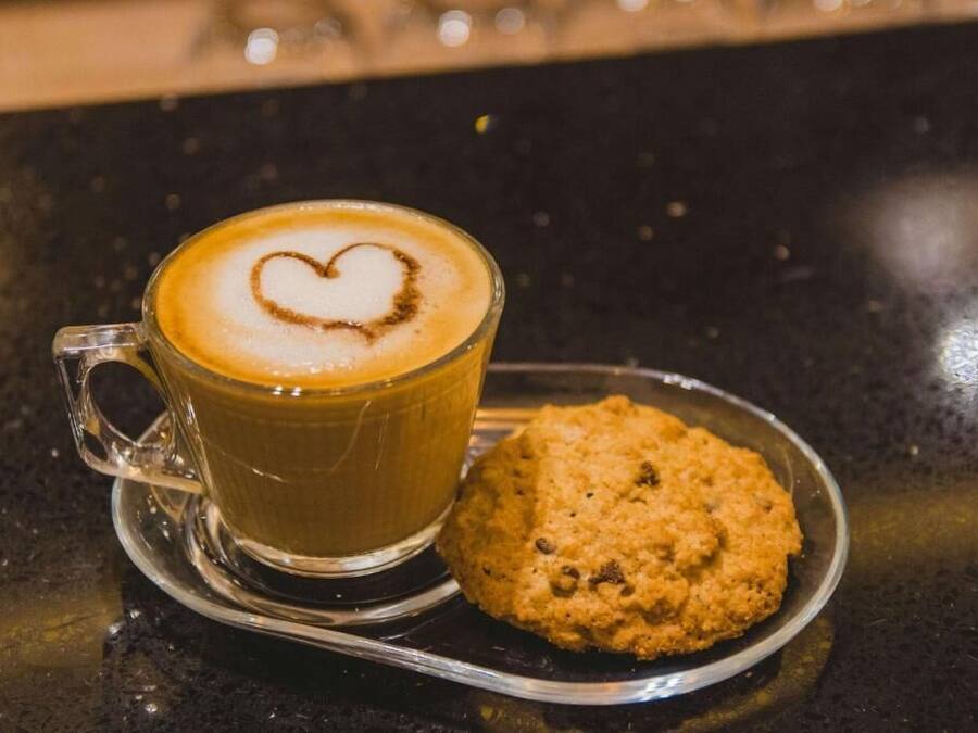 Una taza de café de vidrio transparente con un dibujo de corazón en la espuma, sobre un platito, acompañada de una galleta en un plato pequeño.