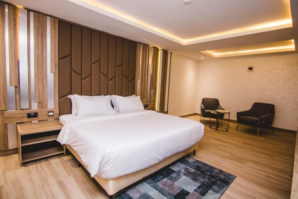 A modern hotel room with a large bed, a wooden headboard, a small desk, and two chairs.
