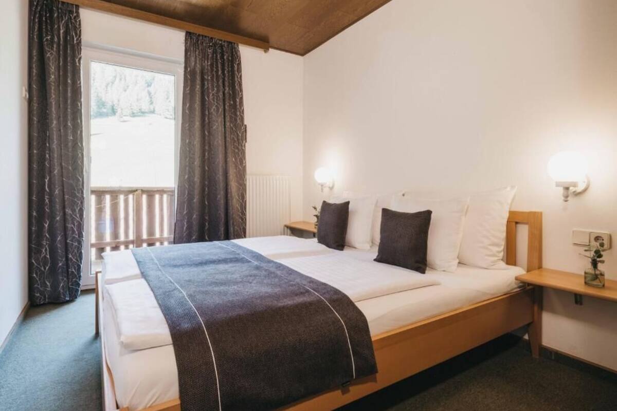 A bedroom with a bed, two bedside tables, a nightstand, a window with curtains, and a view of the outdoors.