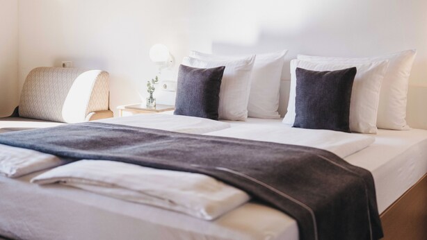 Una cama bien tendida con almohadas blancas y oscuras, una lámpara de noche y un pequeño jarrón con una planta sobre una mesita de noche.