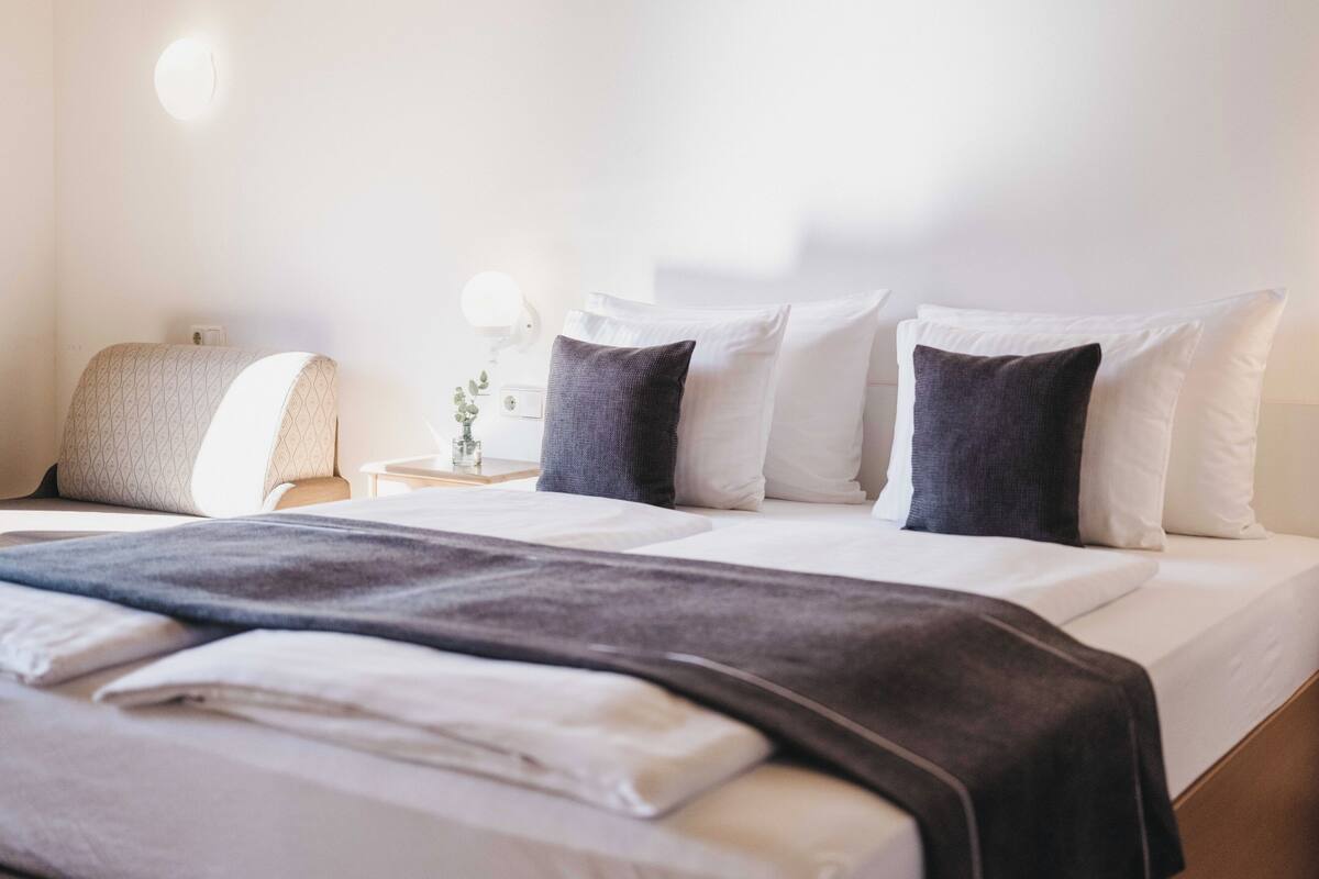 A neatly made bed with white and dark pillows, a bedside lamp, and a small vase with a plant on a bedside table.
