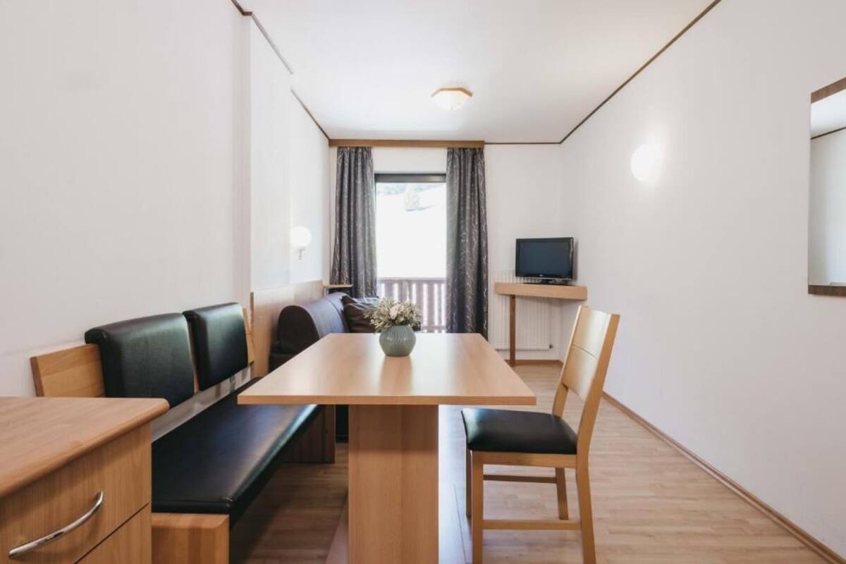A compact room with a wooden dining table, a bench, a single chair, a small TV on a stand, and a window with curtains.
