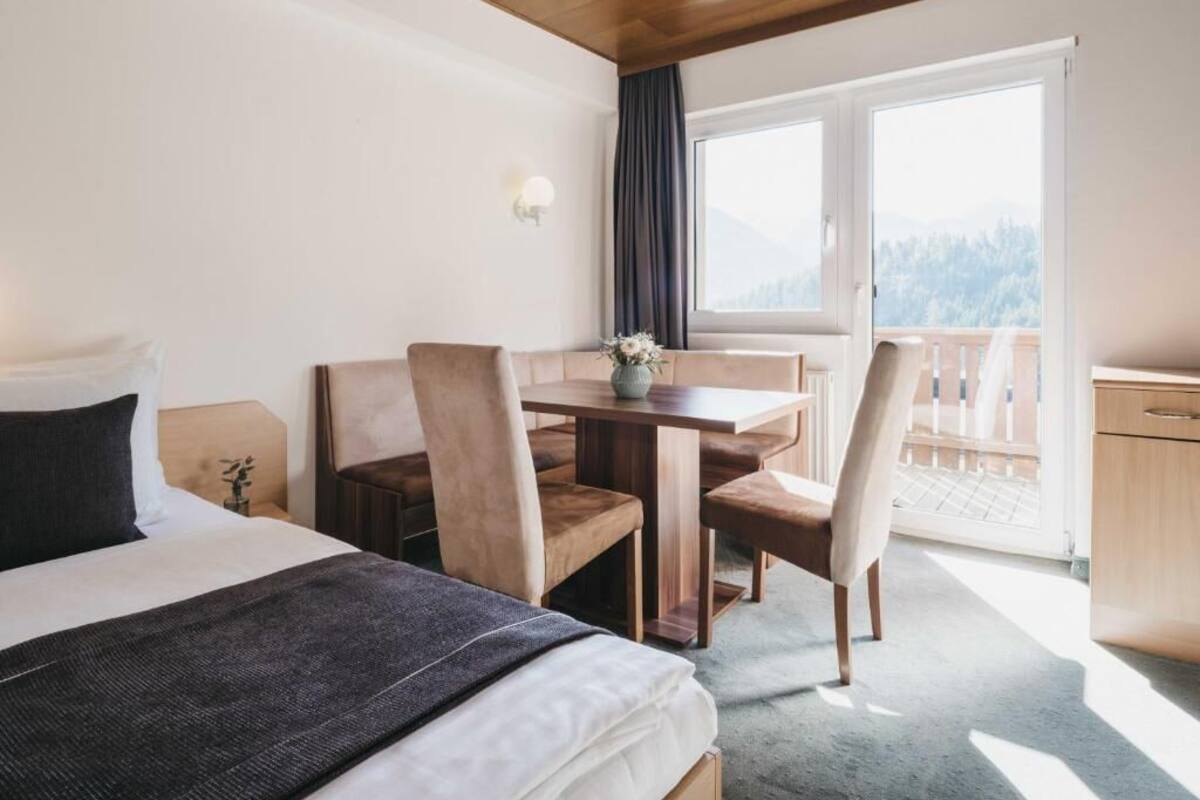 A hotel room with a bed, a dining table with chairs, a small kitchen area, and a balcony with a view of the mountains.