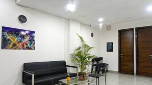 Un área de espera con un sofá negro, una mesa de centro de vidrio y una planta en maceta. Se observan dos puertas de madera y un reloj en la pared.
