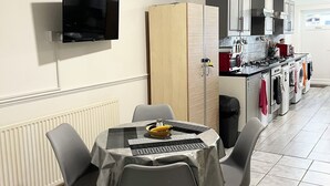 A dining area with a table and chairs, a television mounted on the wall, and a kitchen in the background. - Studio Apartment (Romford)
