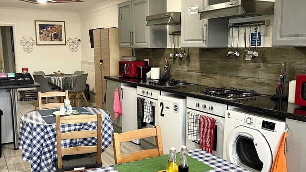 A compact kitchen with a stove, microwave, and a built-in washing machine. There is a dining area with checkered tablecloths and chairs. - Studio Apartment (Romford)