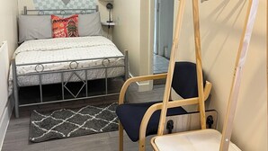 A compact bedroom with a bed, a chair, a small table, and a hanging clothes rack. - Studio Apartment (Romford)