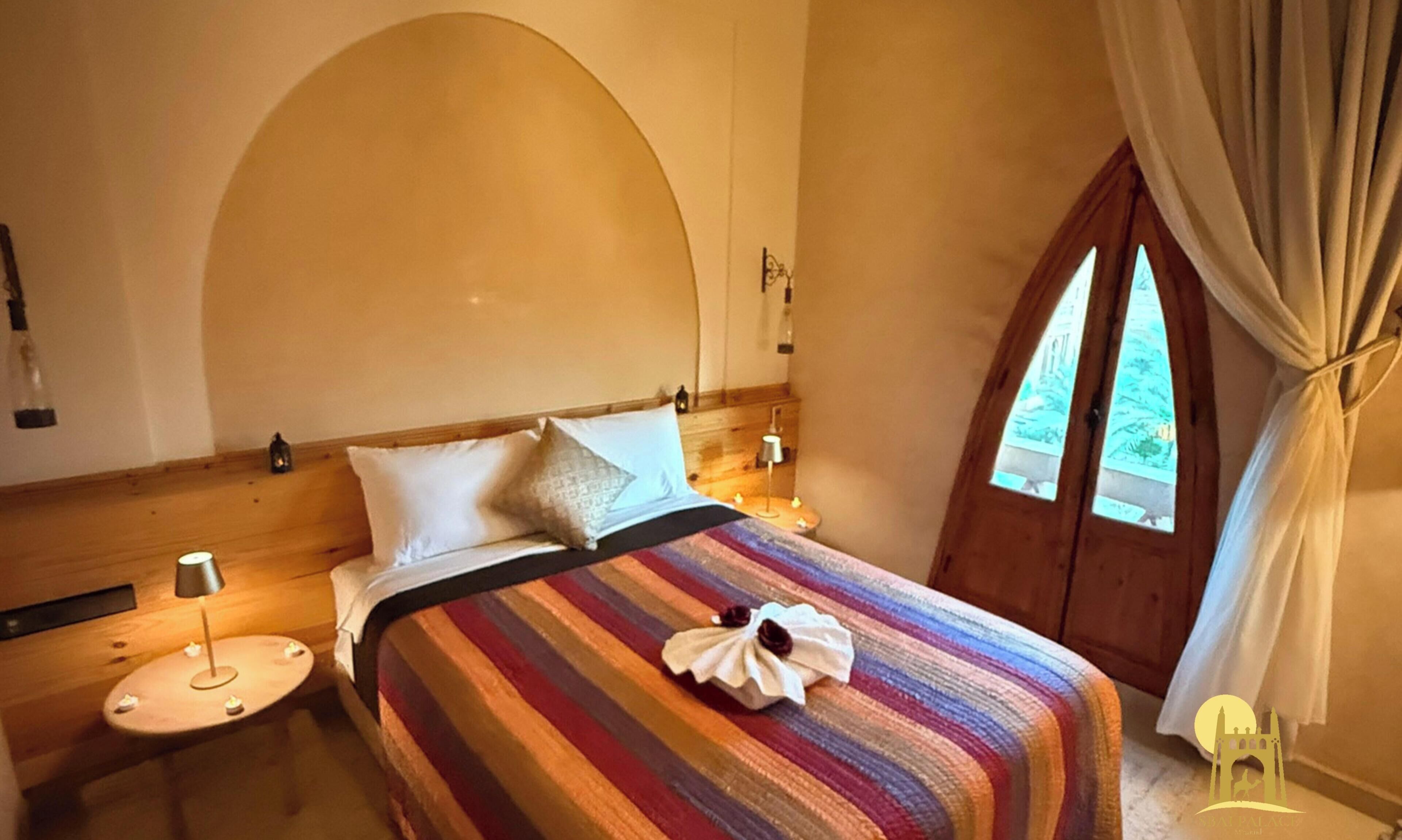 A bedroom with a wooden headboard, a striped bedspread, two bedside tables with lamps, and a window with curtains.