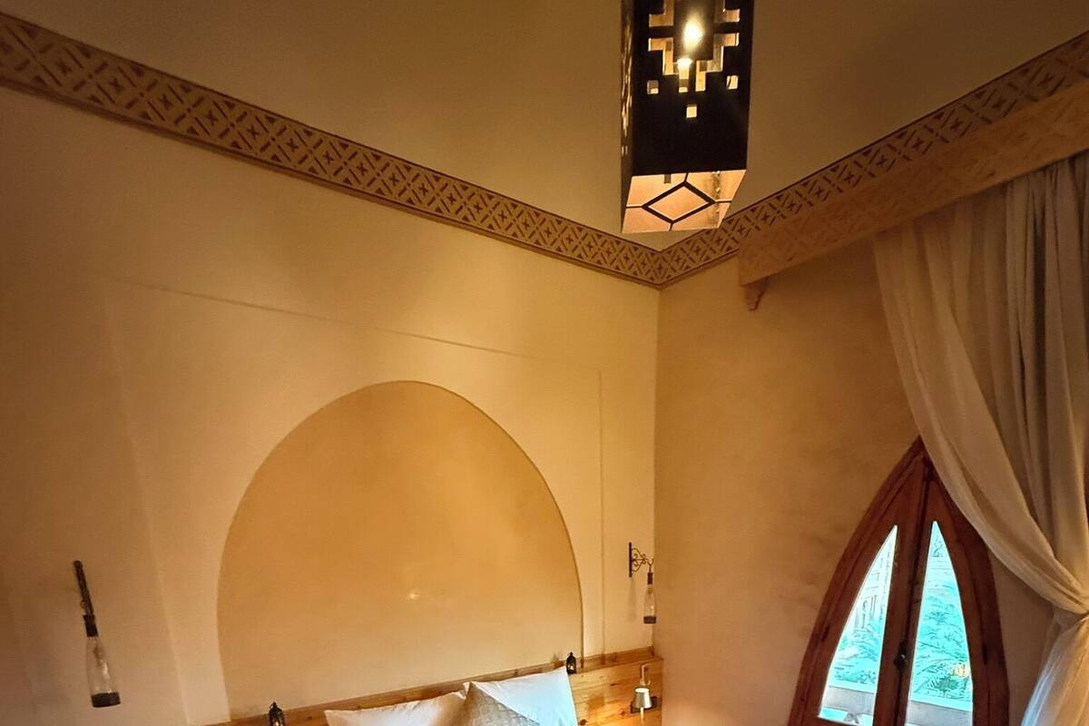 Une chambre d’hôtel avec un grand lit, une tête de lit en bois, une table de chevet avec une lampe et une fenêtre avec des rideaux.