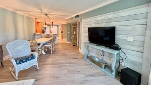 A living room with a television on a wooden stand, a dining area with chairs and a table, and a kitchen in the background. - Tropical getaway awaits!!! (Saint Augustine)