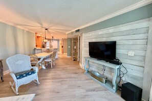 A living room with a television on a wooden stand, a dining area with chairs and a table, and a kitchen in the background. - Tropical getaway awaits!!! (Saint Augustine)