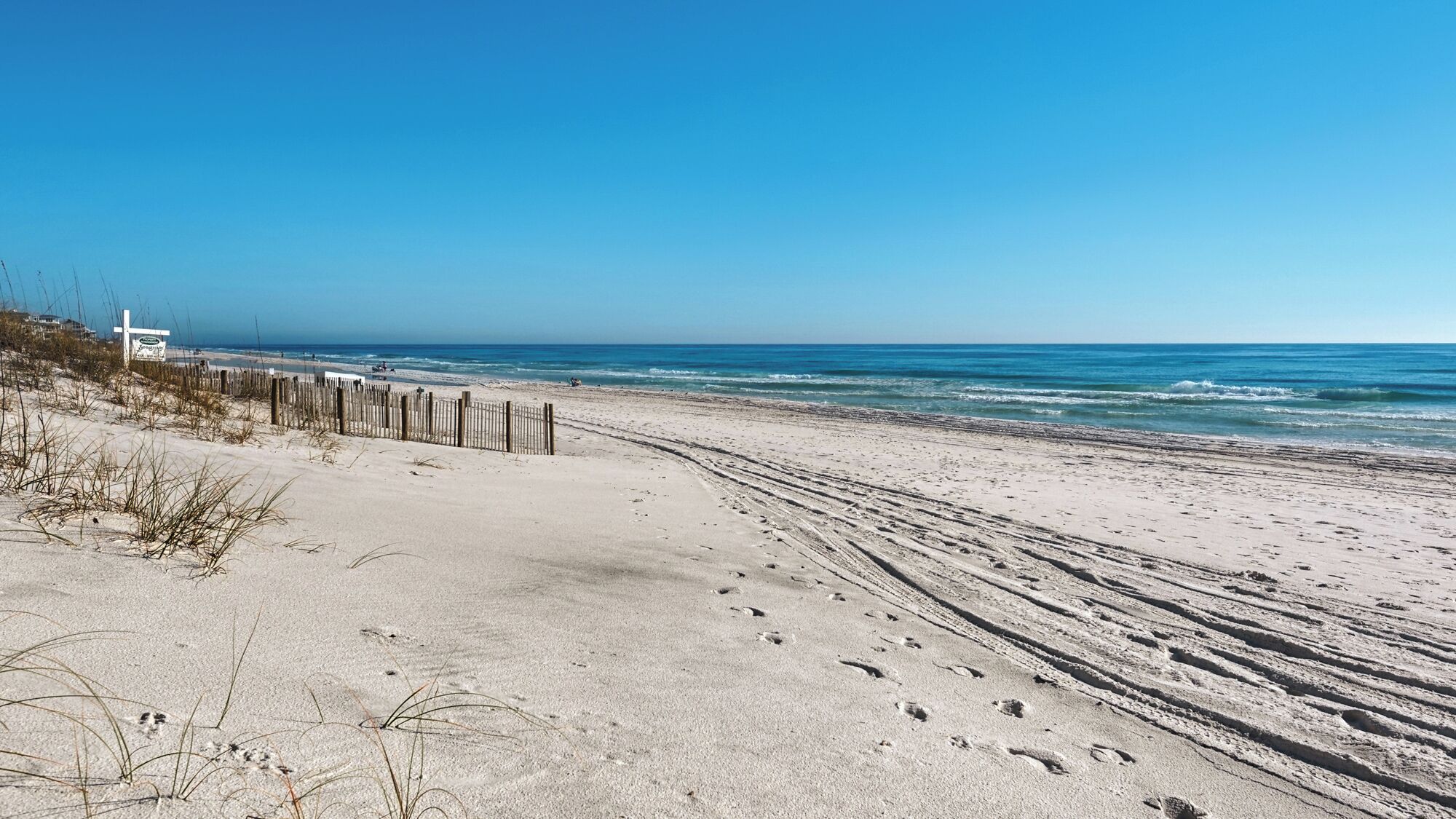 Ein Sandstrand mit Fußspuren, die zum Wasser führen, eine Holzlattenveranda und ein Schild im Hintergrund.
