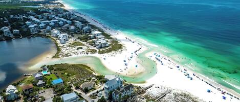 An aerial view of a coastal area with a beach, clear turquoise waters, and a mix of residential and commercial buildings.