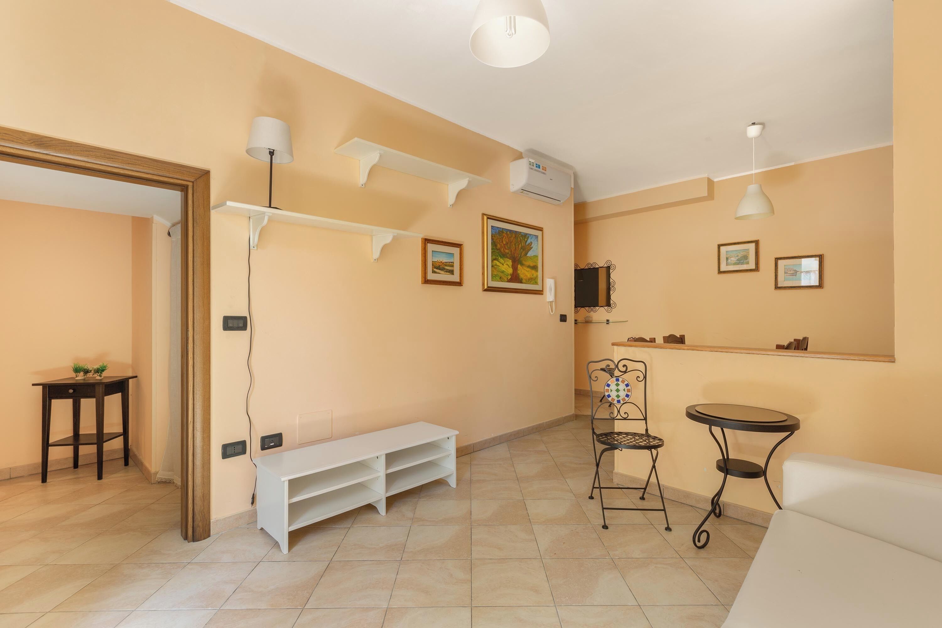 A room with a white bench, a small table, a chair, and a round table. There are wall-mounted shelves and a ceiling fan.