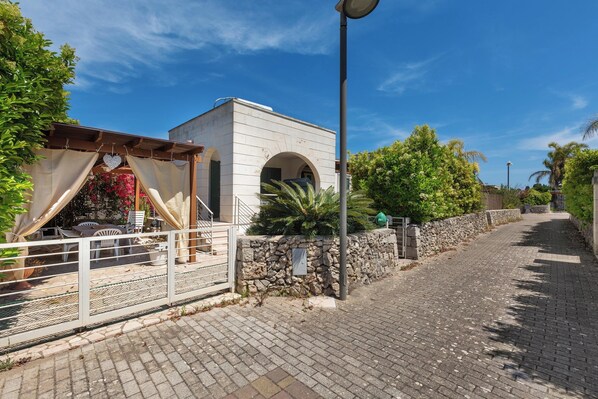 A paved pathway leading to a white building with an arched entrance, surrounded by greenery and a stone wall. - 3945 Villa Ko by Barbarhouse (Otranto)