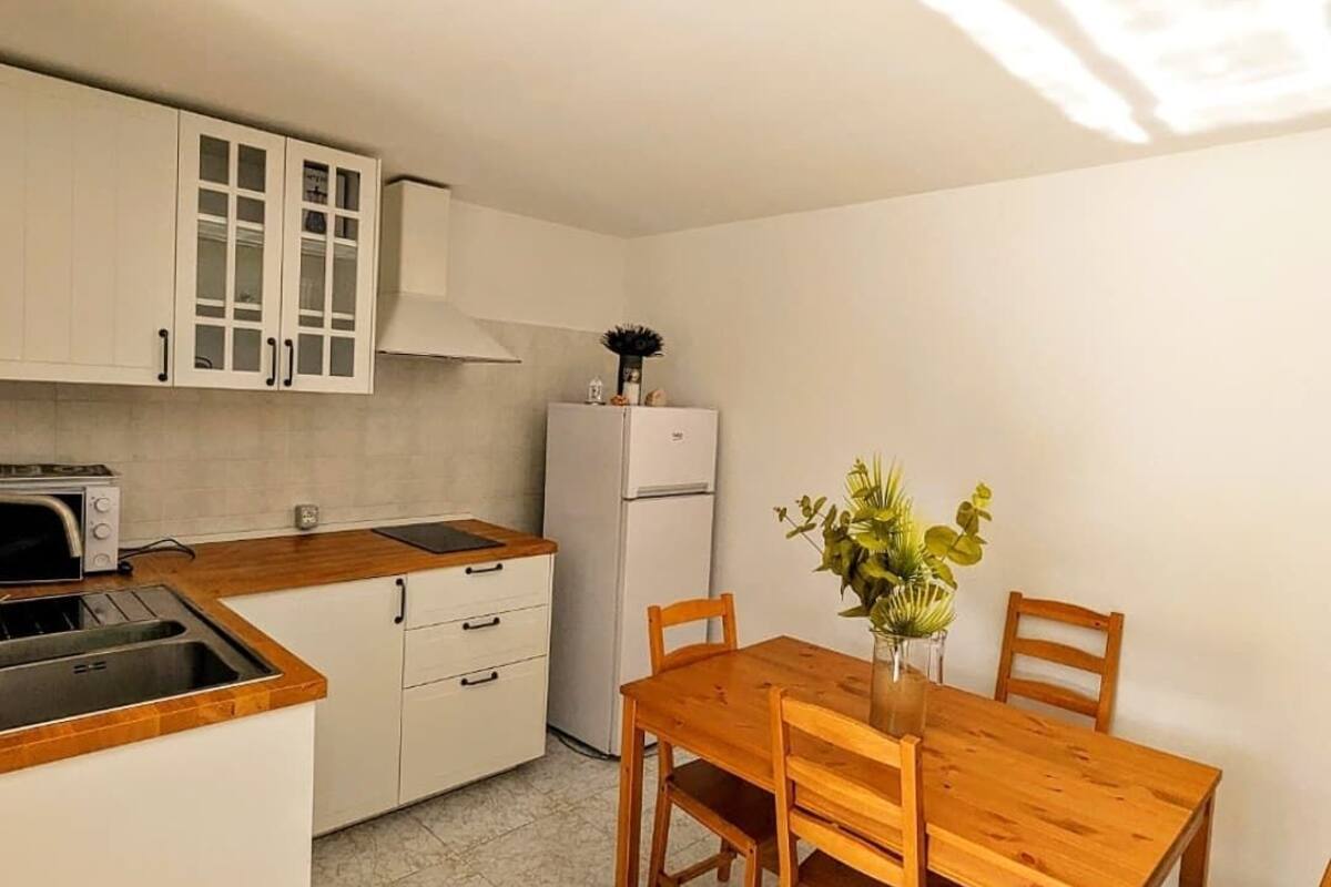 Une cuisine compacte avec des meubles blancs, un four encastré et un réfrigérateur. On y trouve une table à manger en bois, des chaises et un vase de fleurs.