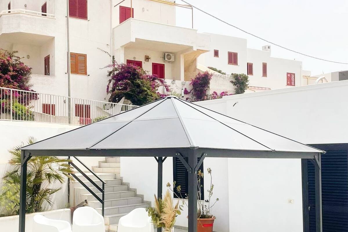 Un espace extérieur aménagé avec une zone de repos couverte, comprenant une table et des chaises, un escalier, et un bâtiment blanc avec des balcons en arrière-plan.