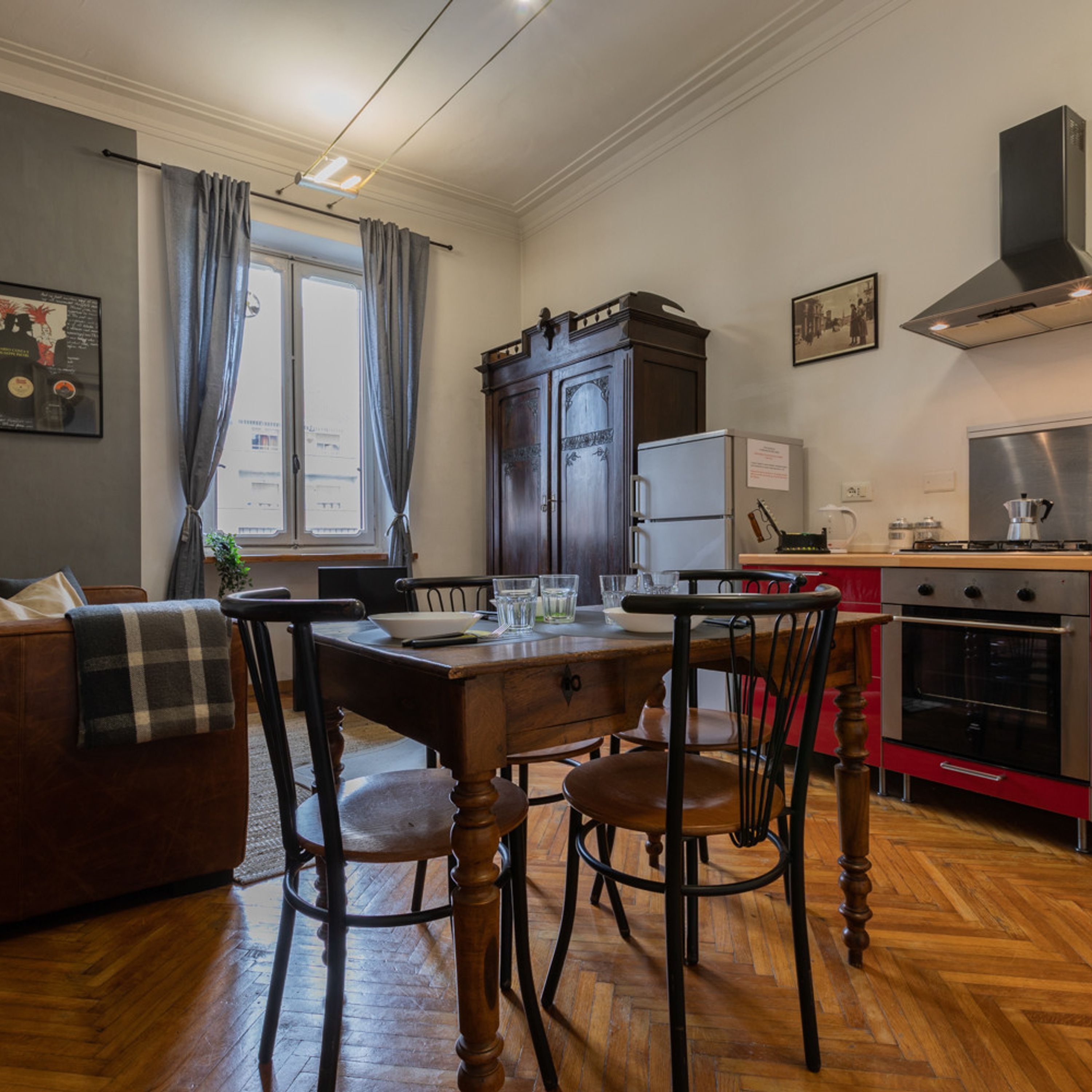 A cozy kitchen with wooden cabinets, a dining table, and chairs. There is a refrigerator, a stove, and a window with curtains.