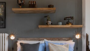 Una cama con edredón blanco, dos almohadas y una manta a rayas. Encima de la cama hay dos estantes de madera con varios objetos.
