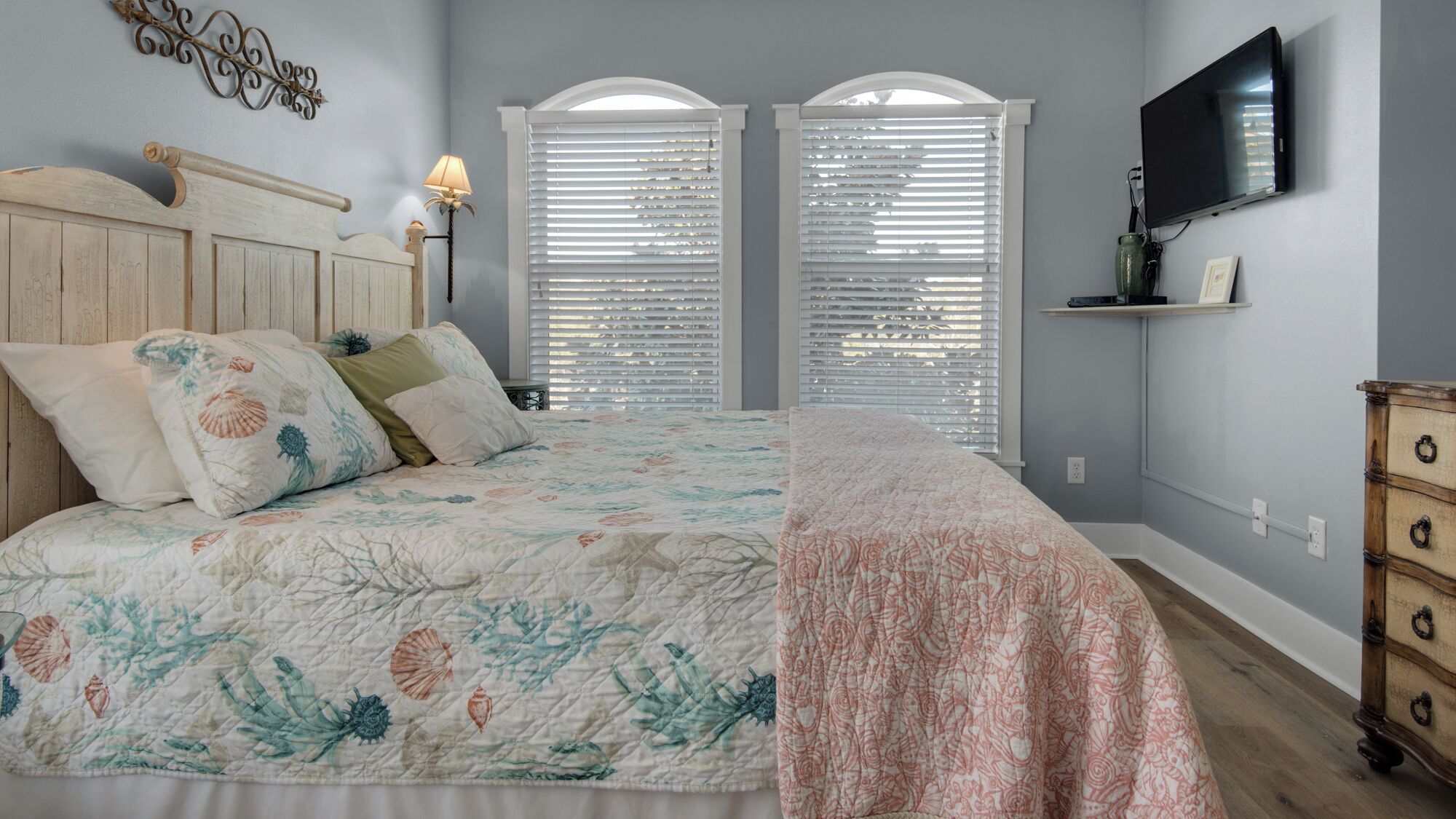 A bedroom with a bed, two windows with blinds, a TV mounted on the wall, and a dresser.