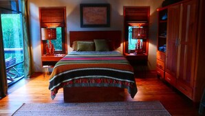 A bedroom with a large bed, wooden flooring, a nightstand with a lamp, a wardrobe, and a window with blinds.