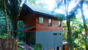 A two-story wooden house with a grey lower level, surrounded by lush greenery and a small garden.