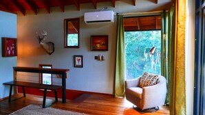 A cozy room with a wooden ceiling, a deer head mounted on the wall, a dining table with chairs, a sofa with a patterned cushion, and a window with curtains.