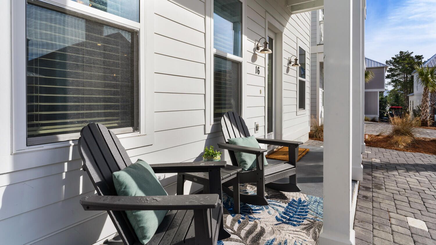Une véranda avec deux fauteuils Adirondack et une petite table, une maison avec un revêtement blanc et le chiffre « 16 » sur la fenêtre.