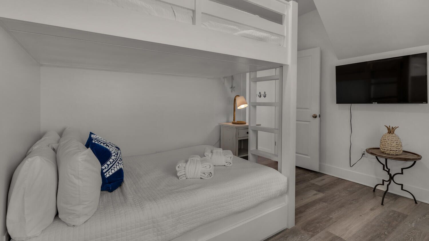 Une chambre avec un lit superposé, une table de chevet avec une lampe, un téléviseur fixé au mur et une petite table ronde décorée d’un ananas.
