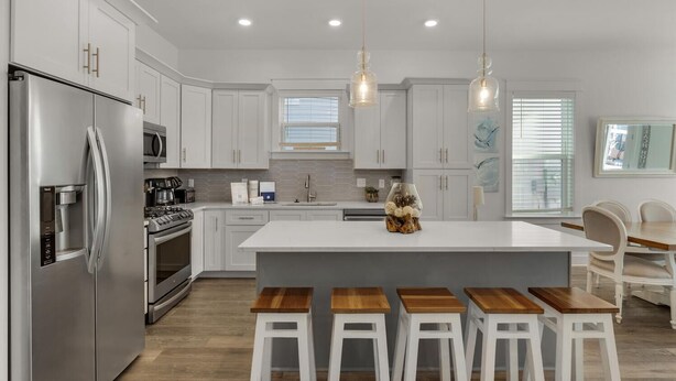 Una cocina moderna con armarios blancos, electrodomésticos de acero inoxidable, una isla central con taburetes e iluminación colgante.