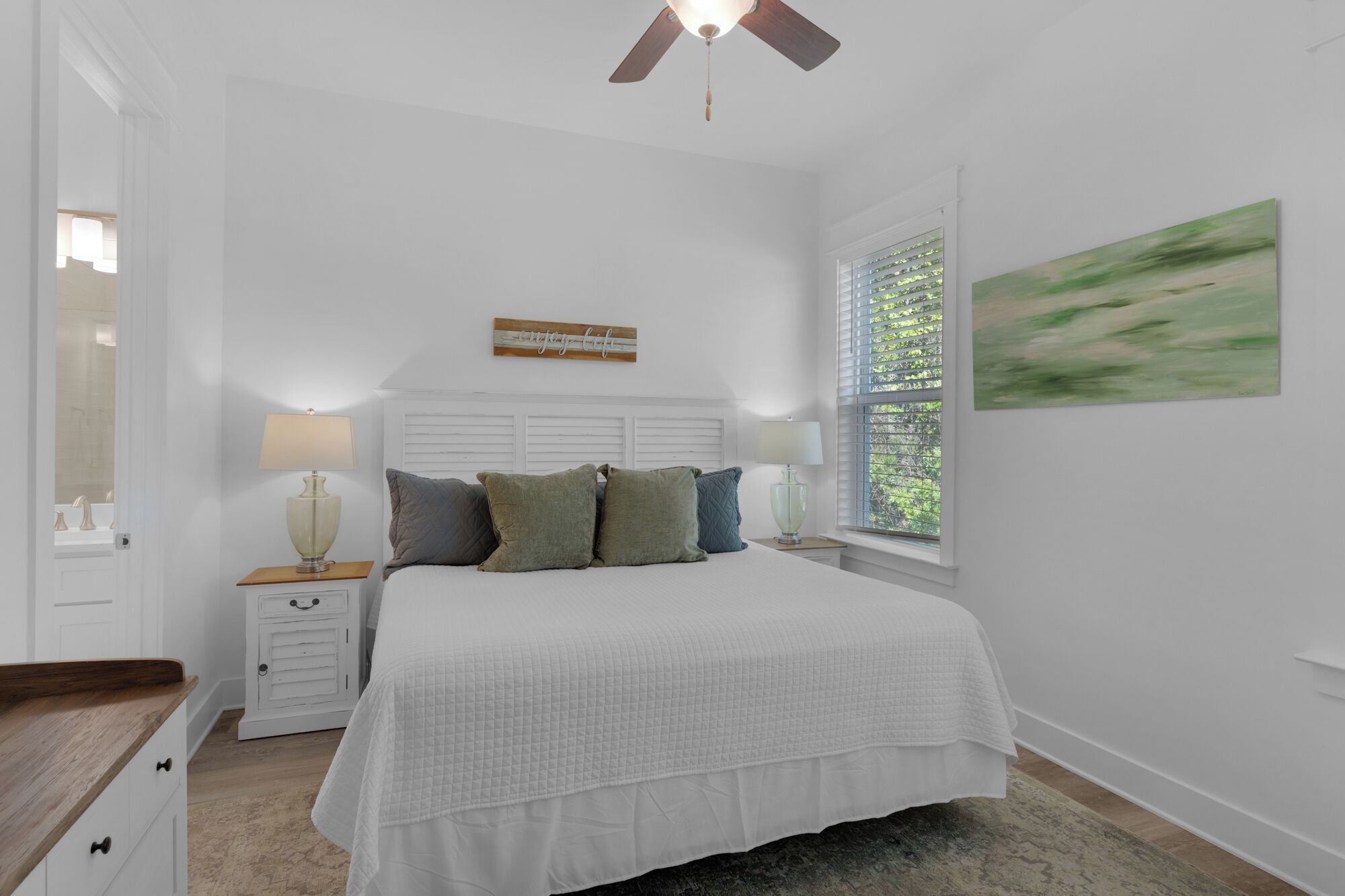 Une chambre à coucher comprenant un lit, deux tables de chevet, un ventilateur de plafond, une fenêtre avec des stores et un tableau accroché au mur.
