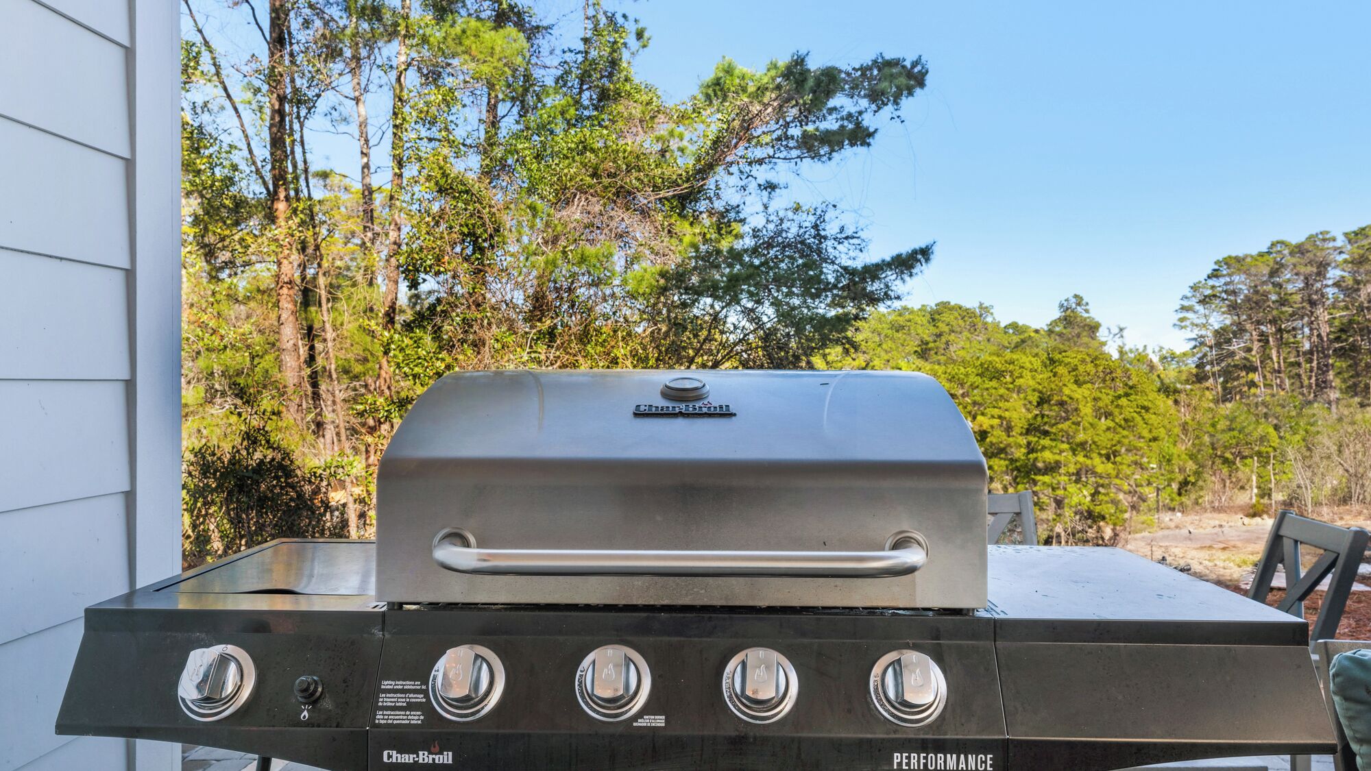 A Char-Broil gas grill with four burners, a control panel, and a lid, set against a backdrop of trees and a house.