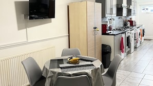 A dining area with a table and chairs, a television mounted on the wall, and a kitchen in the background. - King Studio (Romford)