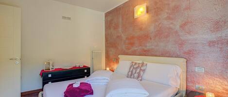 Una cama bien hecha con sábanas blancas, una toalla roja y un cojín decorativo. Se ve una mesita de noche con una lámpara y un pequeño adorno. La habitación tiene suelo de madera y una pared con textura.