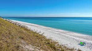 En strand med en trätrappa som leder ner till vattnet, kristallklart blått vatten och en sandstrand.