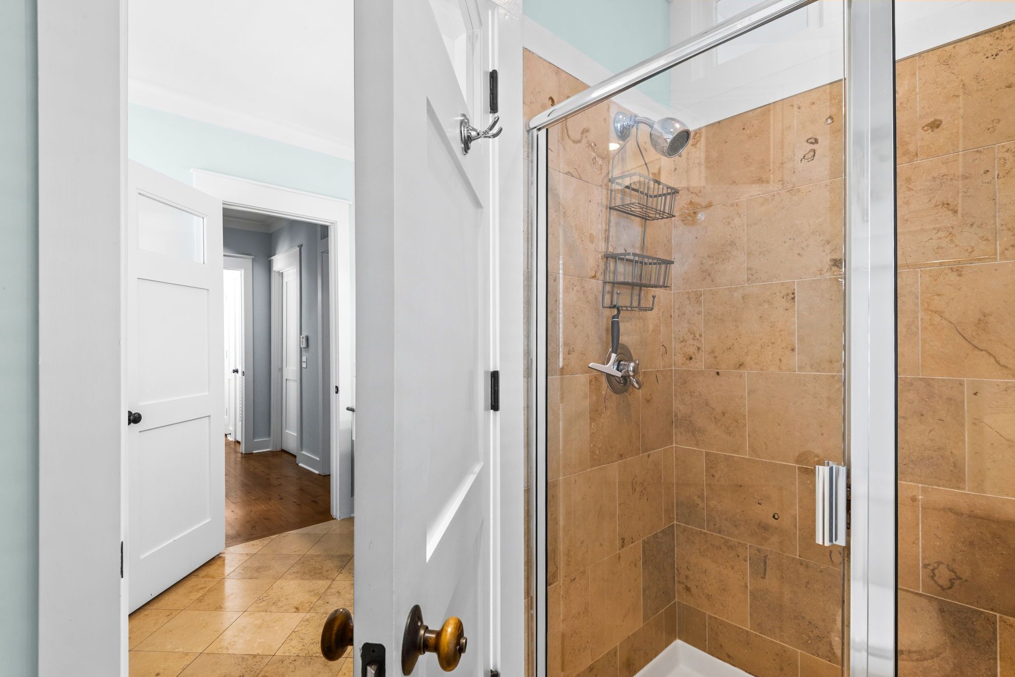 A modern bathroom with a glass shower enclosure, a white door, and a hallway leading to other rooms.