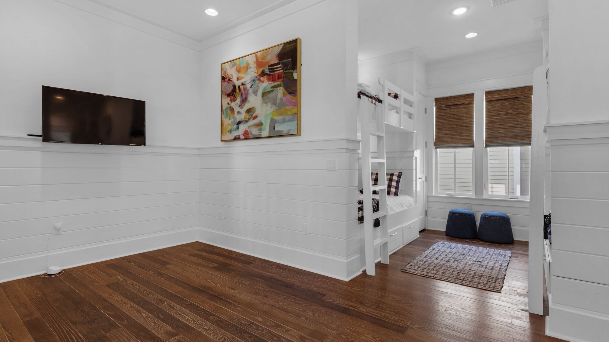 A room with white walls, wooden flooring, a mounted TV, a colorful abstract painting, a built-in bench with storage, and a window with blinds.