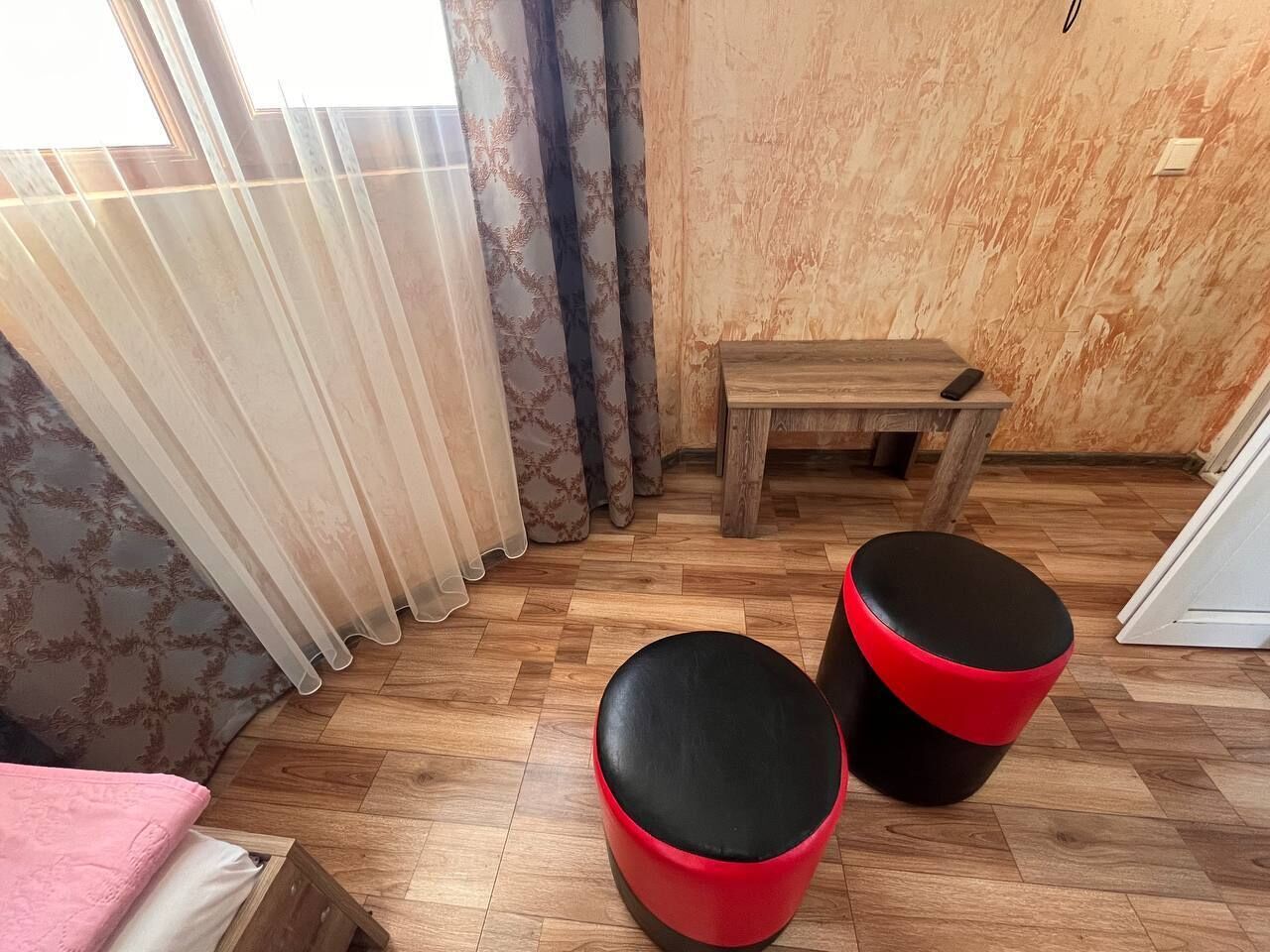 A room with a wooden floor, a bed with a pink blanket, two red and black stools, a wooden bench, and a window with sheer curtains.