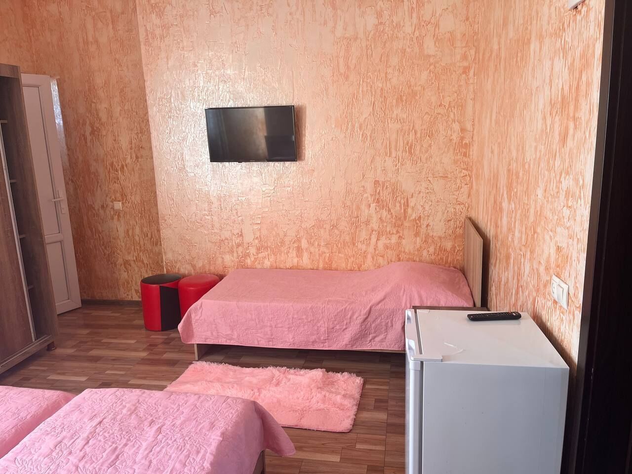 Una habitación pequeña con dos camas, un televisor montado en la pared, un refrigerador pequeño y piso de madera.