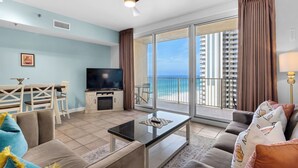 A modern living room with a flat-screen TV, a glass coffee table, and a sofa. There is a balcony with a view of the ocean and buildings. - Amazing Gulf Views! Spa, Pool, Tiki Bar and More! (Panama City Beach)