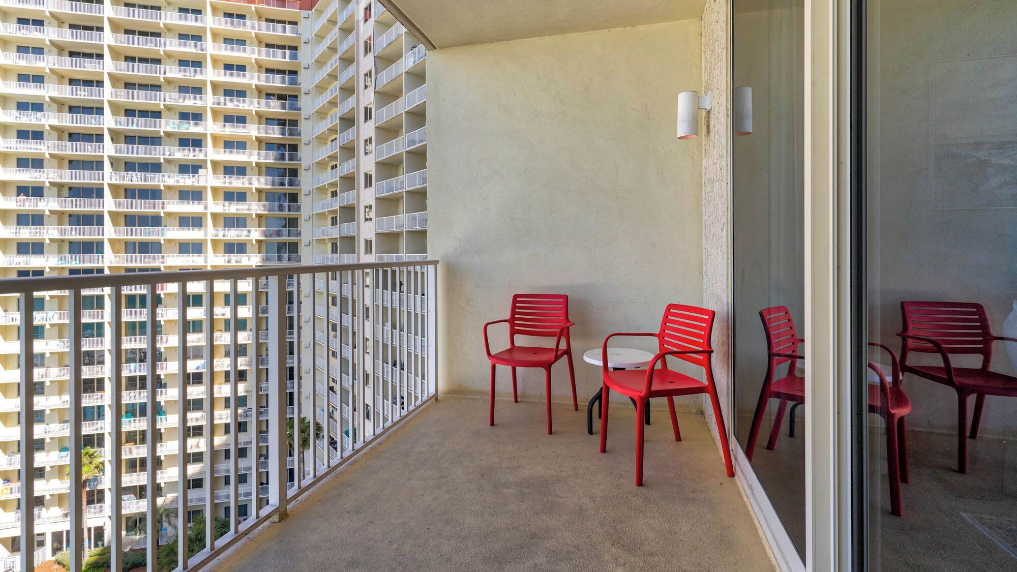 Een balkon met uitzicht op een hoog gebouw, met een glazen deur, een klein rond tafeltje en vier rode stoelen.