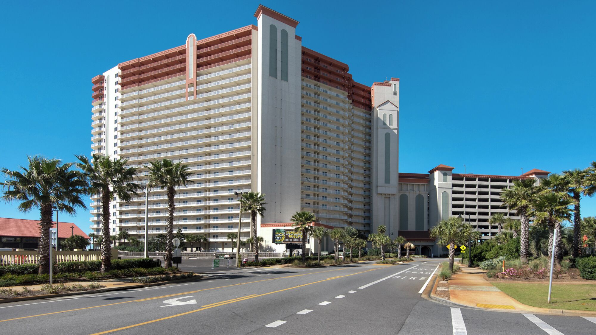 Un grand hôtel à étages, doté d’une tour centrale et d’un parking agrémenté de palmiers.